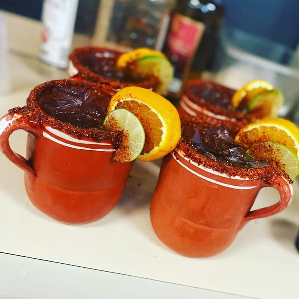 Four Mexican micheladas in red clay mugs, garnished with lime and orange slices and chili powder.