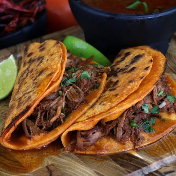 Three birria tacos with shredded meat, cilantro, and lime wedges on a wooden board.