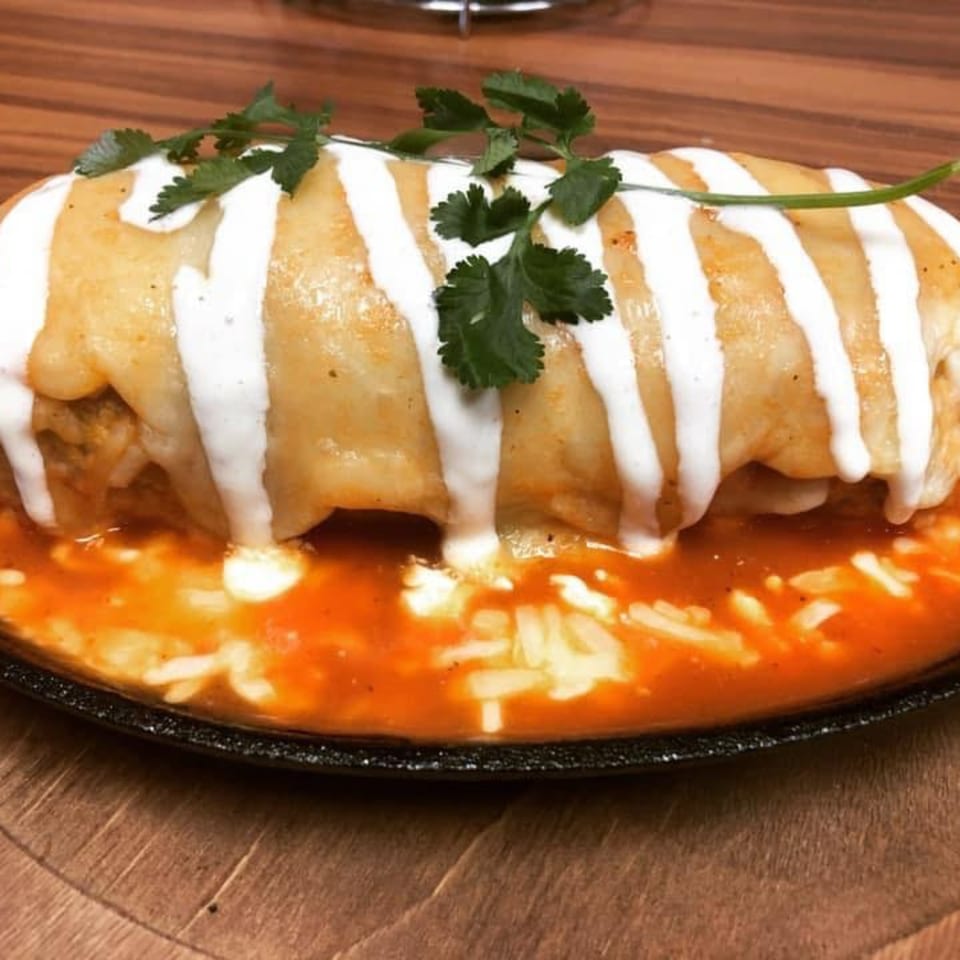 A large, smothered burrito with white sauce stripes and cilantro, sitting in a pool of red sauce.