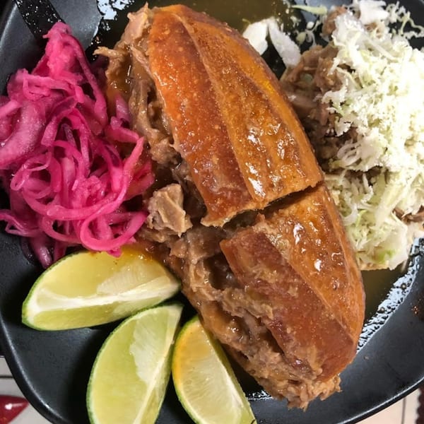 A torta ahogada with pickled red onions, shredded cabbage, cheese, and lime wedges on a black plate.