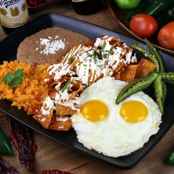 A black plate of chilaquiles with two fried eggs, rice, and refried beans.
