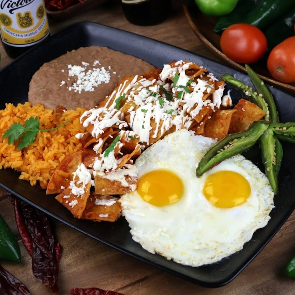 A black plate of chilaquiles with two fried eggs, rice, and refried beans.