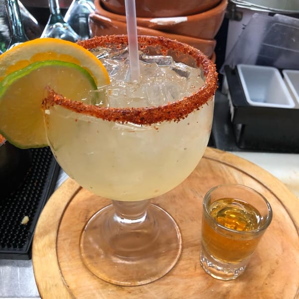 Margarita with a chili-lime rim, lemon and lime slices, and a shot of tequila on a wooden tray.