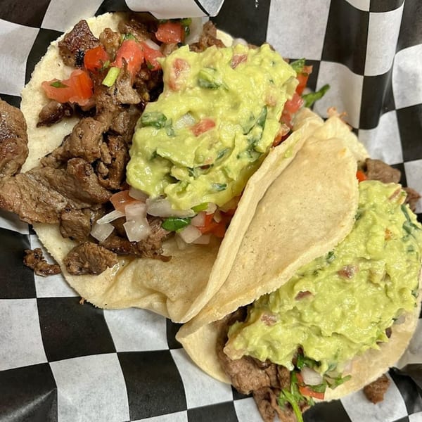 Two carne asada tacos with guacamole and pico de gallo on checkered paper.
